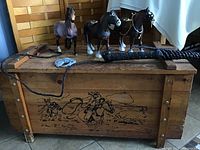 Wood western storage chest with faded painted artwork, three plastic horse figures, metal horse wall hanging, and coiled whip displayed on and around the chest