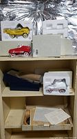 Pedal car models arranged in white styrofoam packaging on wooden shelf including yellow and red pedal cars and additional kits in boxes below.