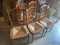 Five wooden chairs with wicker seats arranged in a row in a basement. Two larger captain's chairs with armrests in the front and three smaller spindle back chairs behind them. Some visible wear on wood.