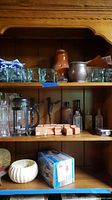 Shelf with clear paneled glass votive holders in large and medium sizes, glass bottles in various shapes, a Bodum French Press, terra cotta pot feet, and a boxed pasta machine.