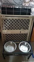 Photo showing metal dog crate with plastic gate and tray behind dog feeder stand with two stainless steel bowls.