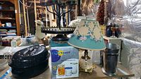 Black speckled roasting pan, metal serving tower, boxed glassware, ceramic cow figurine, brass lamp with floral shade shown on table in basement workshop area