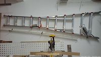 Full view of aluminum ladder stored horizontally on wall with miter saw beneath