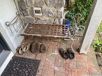 Full view of a weathered metal bench with rusted lattice seat and scrollwork on backrest and armrests, sitting on a brick patio against a stone wall and white siding.