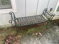 Full view of the metal bench showing lattice seat, scroll armrests, and curved legs placed outside against a siding wall with mossy ground.