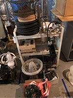 Photo showing a collection of wire bowl/plate holders, a clamp grill light, skewers with wooden handles, and other grilling accessories on a white stand in a basement setting.