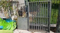 Photo showing metal fence section and gate leaning outdoors. Fence panels have vertical bars and simple metal work; gate has scalloped bottom panel and visible keyhole cutout.