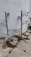 Two metal plant stands with several round platforms for plants, weathered gray finish, tripod bases, shown against a white wall outdoors.