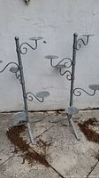 Full view of two metal plant stands with multiple metal circular shelves, weathered condition, outdoors on concrete near white wall.