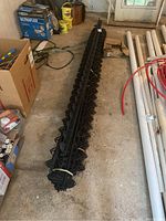 Long bundle of black plastic garden paver liners with interlocking design on a concrete floor, alongside some white pipes and other storage boxes.