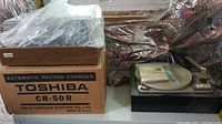 Toshiba CR-50R automatic record changer inside original cardboard box on left, Dual 1006 turntable on right partially covered in plastic, with instruction manual beneath plastic