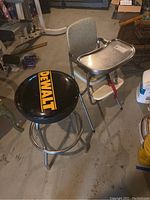 Photo showing the DeWalt branded black metal bar stool next to a metal high chair with tray and safety strap on a concrete floor.