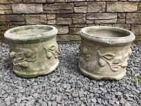 Two side-by-side concrete plant pots with raised leaf patterns, shown from the front on gravel ground with stone wall background.