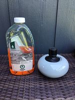 Photo showing full bottle of orange citronella oil next to white ceramic oil burner with black top.
