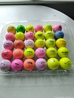 Photo showing 30 coloured golf balls in a plastic tray including colors yellow, pink, orange, green, and blue with brand names visible.