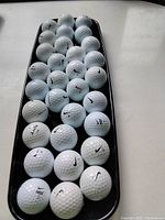 Photo showing 30 white Nike golf balls arranged on a black tray, many with visible Nike swoosh logos and printed numbers, some with "NDX" marking.