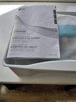 White plastic LG ice maker kit wrapped in foam and blue tape with owner's manual on top.