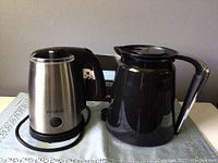 Photo showing the milk frother with stainless steel body and black handle alongside the black plastic double walled carafe with handle and lid.