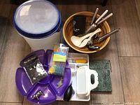 Kitchen items including large plastic storage container with lid, wooden bowl, cooking utensils, purple segmented tray, and marble cheese slicer.