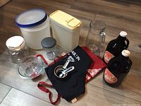 Photo showing all items in the lot including aprons, measuring cups, glass mixing bowl, storage containers, glass juice jug, and growler jugs arranged on floor.