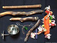 Five natural wood perches, colorful rope and wood bird toys, and a stainless steel food dish with mounting bracket displayed on a black background.