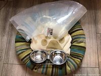 Full view of pet bed with paw print cushion, clear plastic cone collar, stainless bowls at front