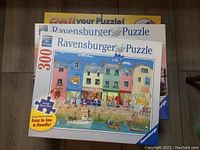 Two boxed Ravensburger puzzles, one labeled 300 pieces with harbor scene, one 1000-piece panorama puzzle showing gondolas in Venice, stacked on floor with box for felt puzzle mat beneath