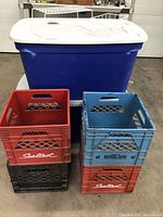 Photo of two large blue storage totes with white lids, stacked milk crates in red, blue, and black in front