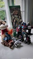 Photo showing all toys on a white surface in front of windows: boxed doll, three stuffed animals, transformer robot toy.
