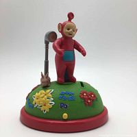 Front view of vintage Po Teletubby piggy bank on a green floral base with rabbit and coin slot.