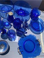Various cobalt blue glassware including bowls, pitcher, small glasses, and serving dishes displayed under bright light showcasing the deep blue color and transparency.