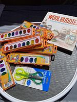 Photo showing multiple Crayola watercolor paint palettes, a pair of kids' scissors in packaging, the white lazy Susan tray, and the craft book "Wool Buddies" arranged on the tray.