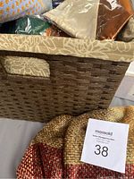 Woven basket filled with various fabric items including chair sashes and chenille blanket along with a boxed bud vase and folded table cover.