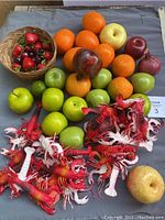 Wide view of all faux food items including apples, oranges, strawberries in small basket, cherries, pear, onions, and red and white faux crawfish arranged on a flat surface.