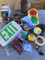 Wide view of all items arranged on a surface showing the exit sign, duct tape rolls, safety ponchos, emergency light, stapler, screws, measuring tape, leather pouch, and flashlight.