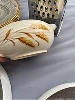 Close-up of wheat-patterned vintage Pyrex small bowl with handle and partial view of metal box grater and gold rim plates.