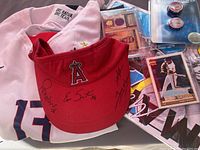 Signed red Angels visor with multiple autographs, white Nike soccer jersey with USA crest and number 13 visible, assorted baseball cards and PSA-graded magazine on table.