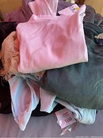 Pile of assorted women's clothing including pink ribbed top, green long sleeve shirt, beige knit, white clothing item, and a lavender puffer jacket underneath