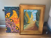 Two original paintings: one framed street scene and one unframed abstract painting, shown side by side on floor against wall.