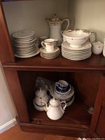 Multiple pieces of white china including plates, cups, saucers, teapot, sugar bowl, and coffee pot arranged inside a wooden cabinet.