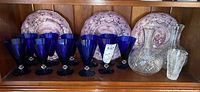Shelf display featuring two clear cut crystal decanters, eleven cobalt blue glasses, and three decorative purple plates.