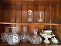 Five decanters and small china plates on white pedestal dish on wooden shelf