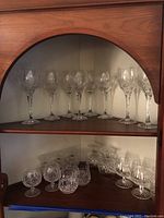Overall view of 21 crystal glasses arranged on two wooden shelves inside a cabinet, showing a variety of clear cut crystal stemware styles.