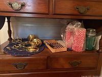 Two bronze-toned candleholders with intricate scroll designs placed on a dark quilted cloth on a wooden surface alongside packages of red and white striped candles and a green jar candle.