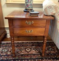 Front view of one wooden end table showing two drawers with brass handles and turned legs.
