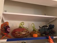 Shelf view showing all seven glass statuettes: millefiori fish, three pumpkins, shark with small fish, spiky fish