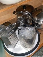 Photo showing metal box grater, metal over-sink colander, some trivets and pots stacked on a white tray