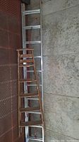 Werner aluminum extension ladder fully extended on left side and wooden ladder placed atop it leaning against a metal wall.