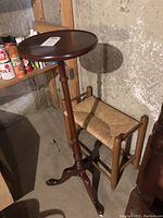 Wooden pedestal plant stand with round top beside small wooden bench with woven wicker seat, shown in corner of room.