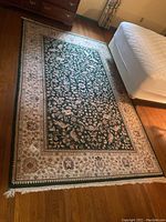 Full view of green and cream floral patterned area rug on hardwood floor with white fringe on two edges. Rug is rectangular and placed near a bed.